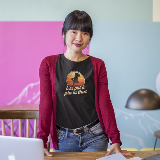 Person standing in an office with a laptop and books on a desk, wearing a black t-shirt with text and graphics.