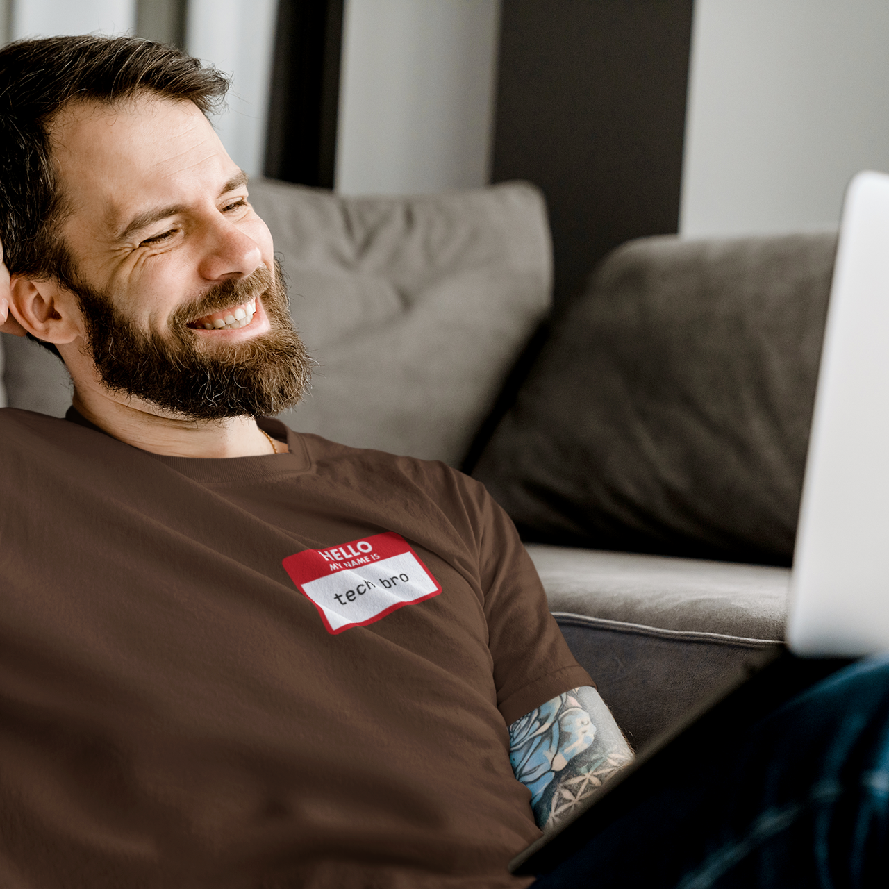 Man with tattoos and a beard wearing a brown t-shirt with a name tag, sitting on a couch.