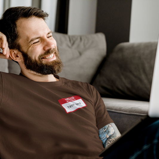 Man with a beard wearing a brown t-shirt with a name tag, sitting on a couch using a laptop.