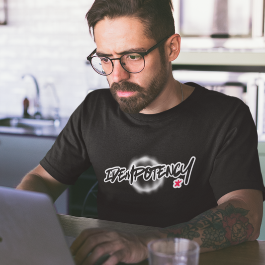 Man wearing a black t-shirt with a logo and text, sitting at a table using a laptop in a kitchen.