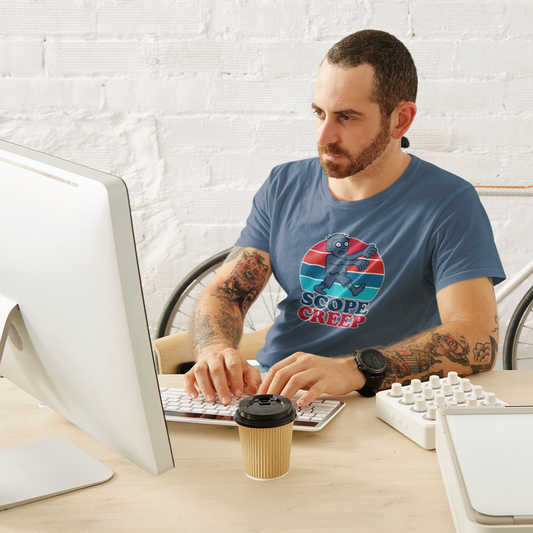 Man sitting at a desk with a computer, wearing a t-shirt with a colorful design, with a bicycle in the background.