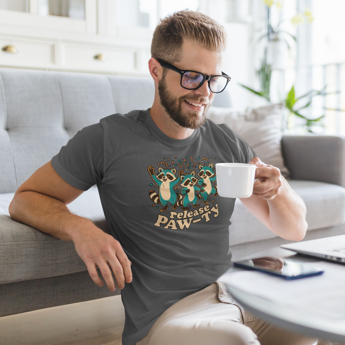 Man sitting on a couch holding a mug, wearing a t-shirt with raccoon graphics and text.