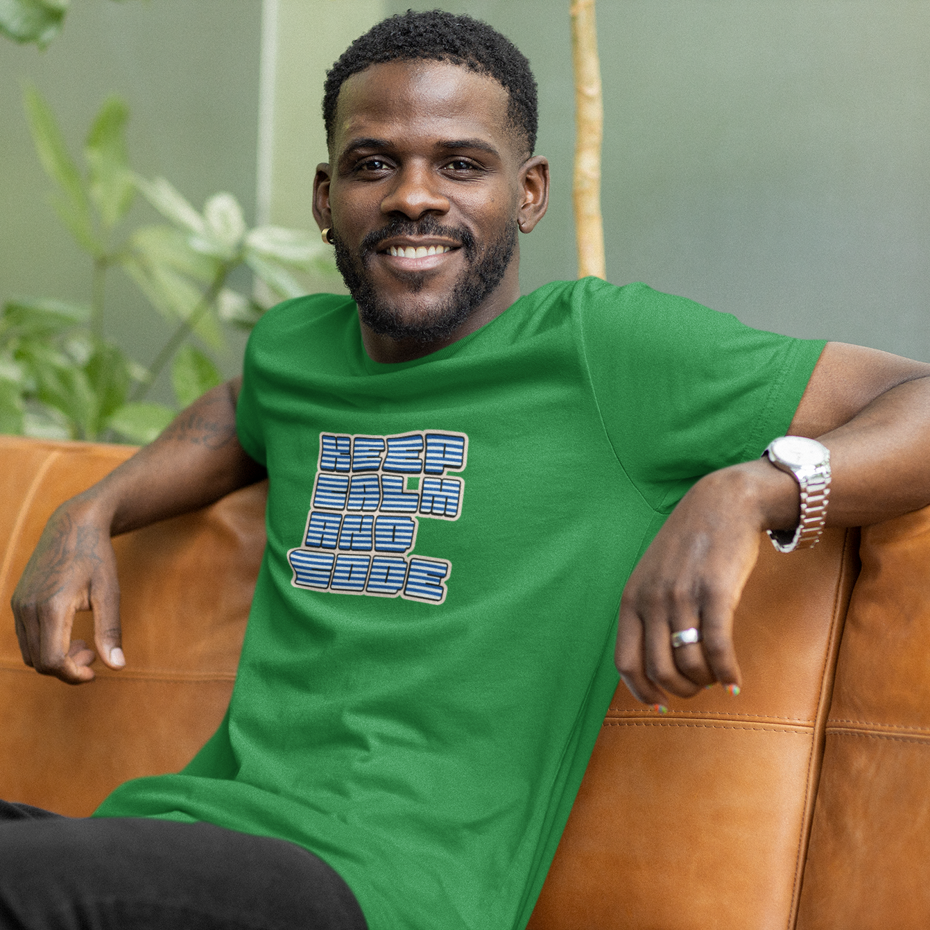 Man wearing a green t-shirt with a graphic design, sitting on a brown couch with a blurred green plant background.