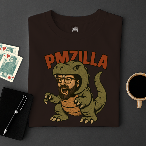 Black t-shirt with a graphic design of a man in a dinosaur costume with 'PMZILLA' text, placed on a dark surface with playing cards and a coffee cup.