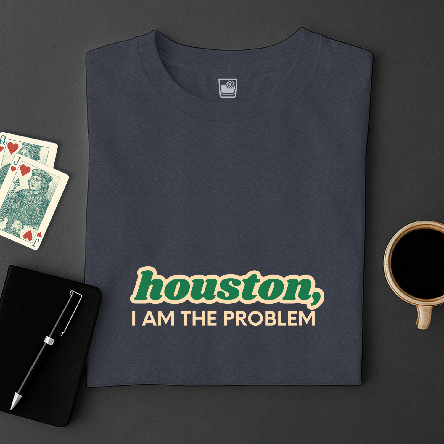 Navy blue t-shirt with 'Houston, I am the problem' text on a dark surface with cards and a cup.