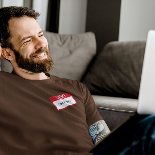 Man with tattoos and a beard wearing a brown t-shirt with a name tag, sitting on a couch.