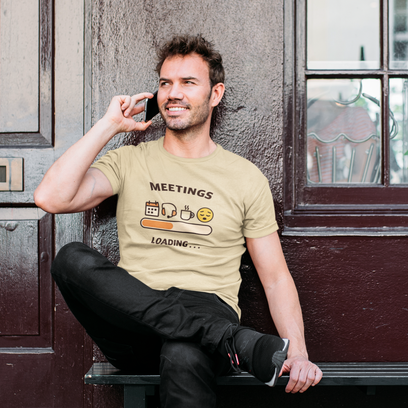 Man wearing a beige t-shirt with 'Meetings Loading' graphic sitting against a door.