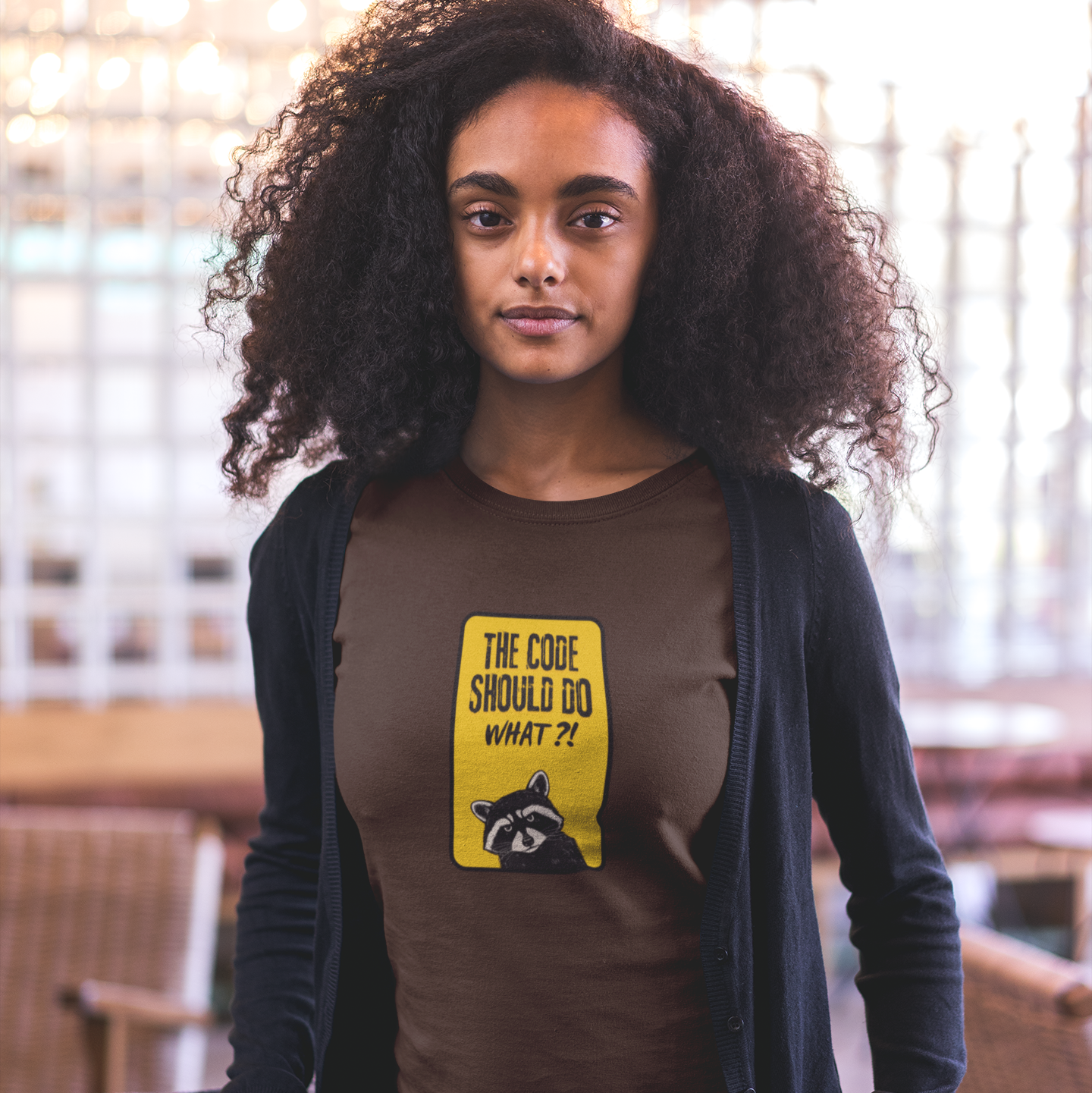 Woman wearing a brown t-shirt with a yellow graphic and text, standing indoors.