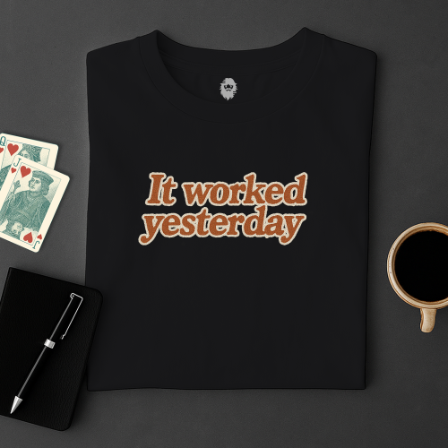 Black t-shirt with 'It worked yesterday' text on a dark surface with cards and a cup.