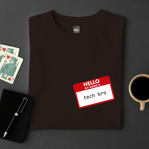 Black t-shirt with 'Hello My Name Is tech bro' tag on a dark surface with cards, pen, and coffee cup.