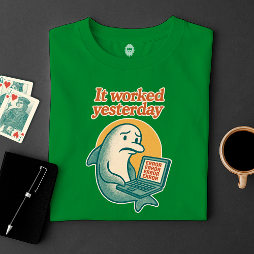 Green t-shirt with a dolphin graphic and text 'It worked yesterday' on a dark surface with cards and a cup.