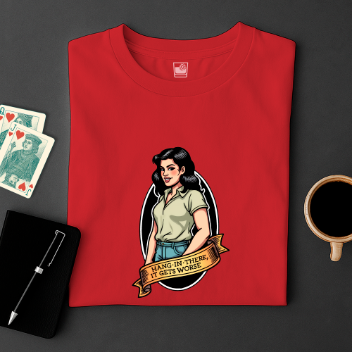 Red t-shirt with graphic design of a woman and text 'Hang in there, it gets worse' on a dark surface with cards and a cup.