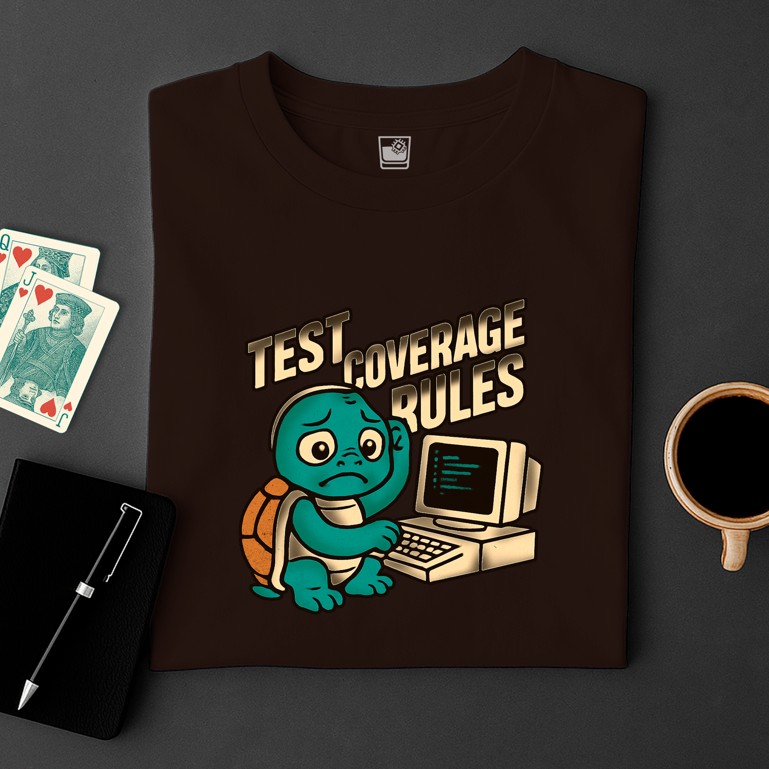 Black t-shirt with a turtle and computer graphic and 'Test Coverage Rules' text, placed on a dark surface with cards and a cup.