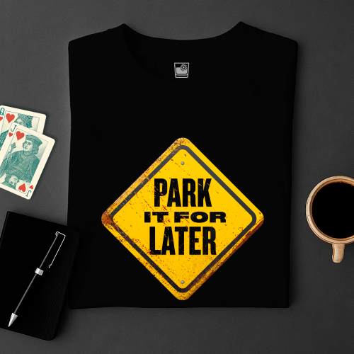 Black t-shirt with yellow 'Park It For Later' sign design on a dark surface with cards and a cup.