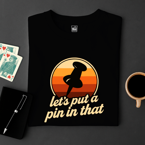 Black t-shirt with a graphic design and text on a dark surface with cards and a cup.