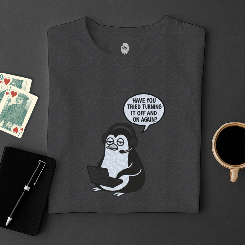 Dark gray t-shirt with a penguin graphic and text, placed on a dark surface with playing cards and a coffee cup.