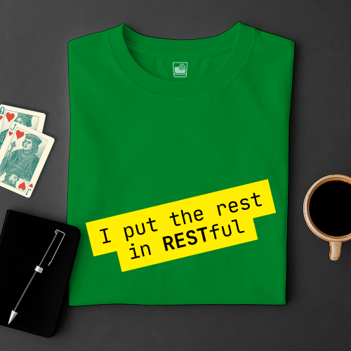 Green t-shirt with yellow text 'I put the rest in RESTFUL' on a dark surface with playing cards and a cup.