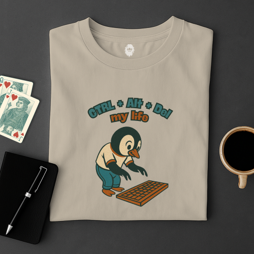 Folded t-shirt with a penguin graphic and text on a dark surface with playing cards and a coffee cup.
