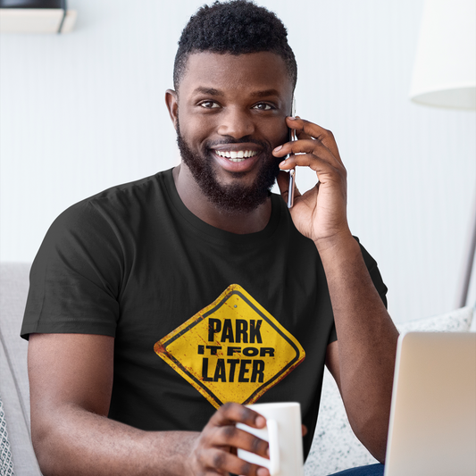 Man sitting on a couch with a laptop, holding a coffee cup, and talking on a phone. He is wearing a black t-shirt with a yellow and black graphic design.