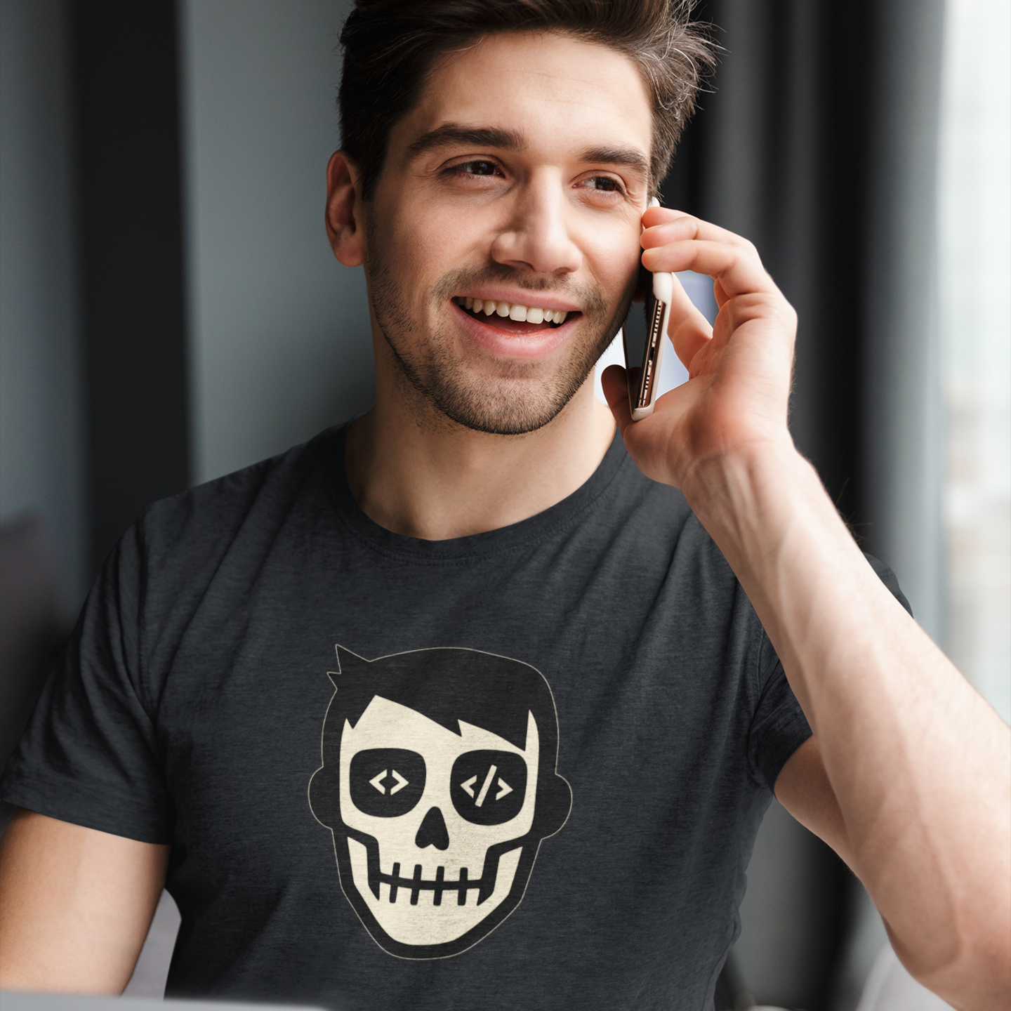 Man talking on a phone with a laptop in front of him, wearing a black t-shirt with a skull and code design.