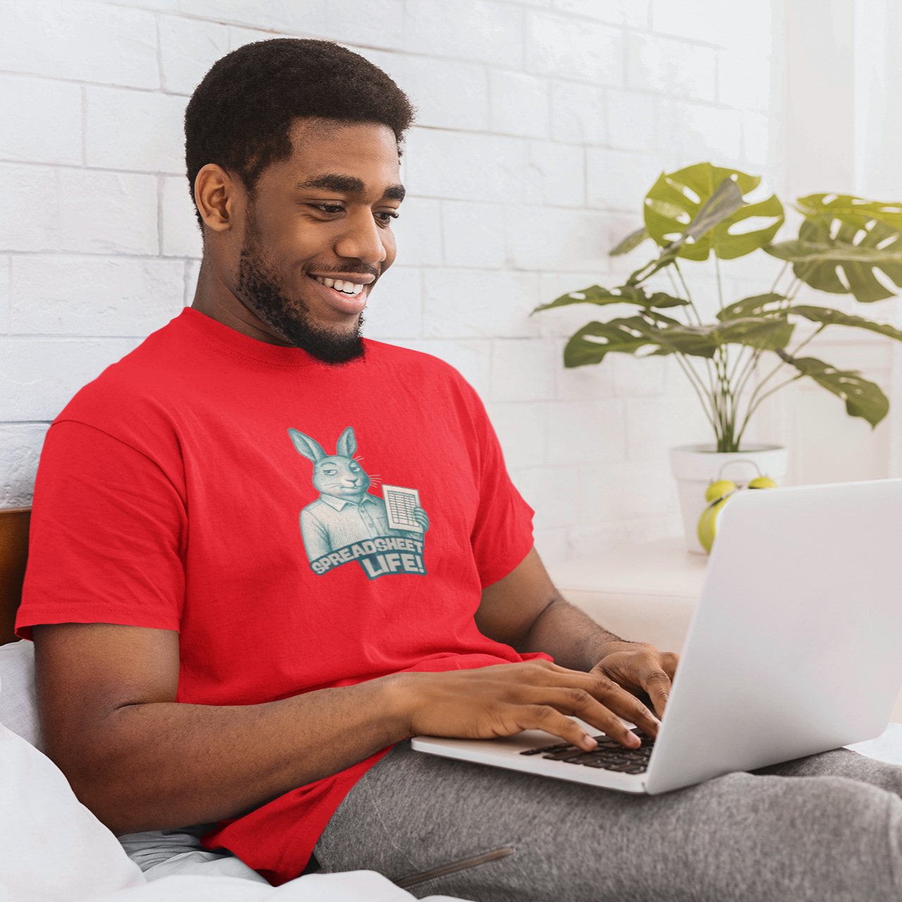 Man in a red shirt using a laptop on a bed with a plant in the background