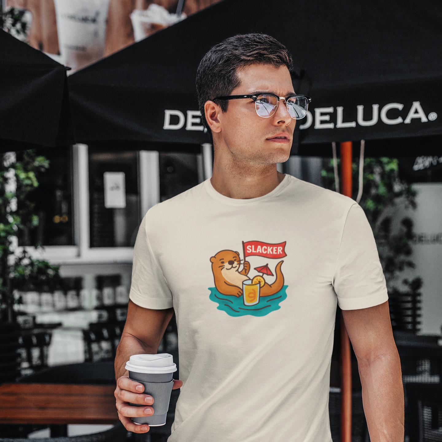 Man wearing a t-shirt with a otter that is a slacker, holding a coffee cup, standing in front of a coffee shop.