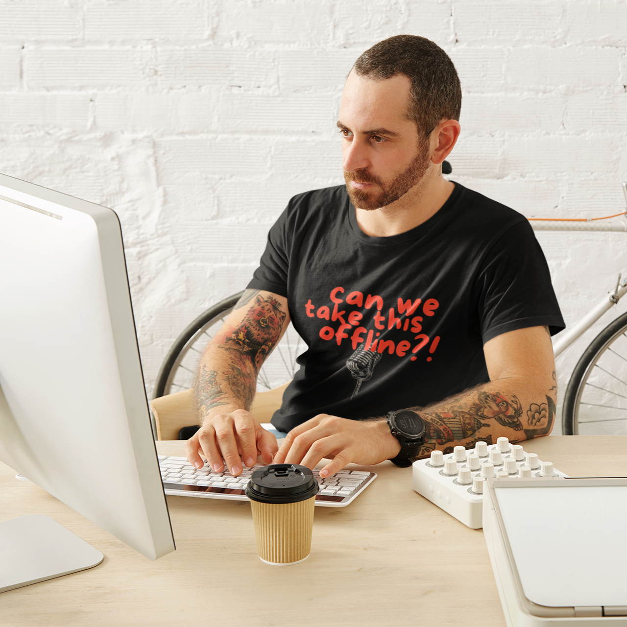 Man sitting at a desk with a computer, wearing a black t-shirt with red text, in an office setting with a bicycle in the background.
