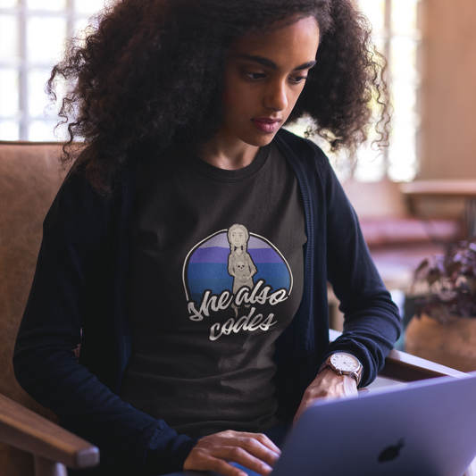 Woman using a laptop wearing a shirt with 'She Also Codes' design.