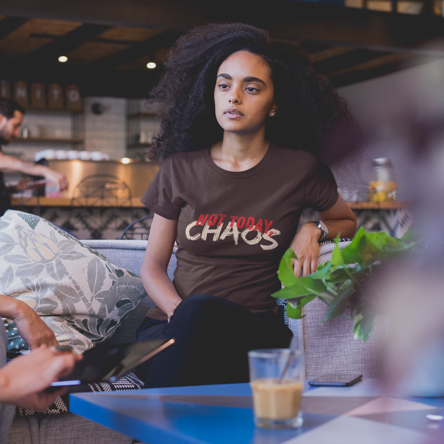 Woman wearing a 'Not Today Chaos' t-shirt sitting on a couch with a plant in a cafe.